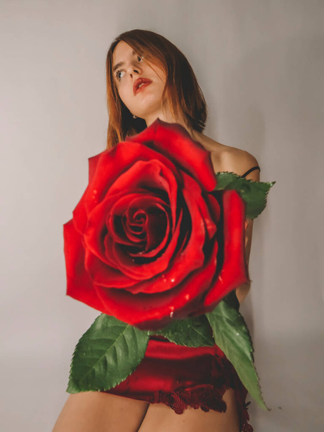 A young brown-haired woman with an attractive appearance in a red negligee with a red rose flower in her hands on a light wall background, close-up, the concept of the holidays of St. Valentine, March 8,signs of attention,romantic dates,female beauty - Fotoğraf, Görsel