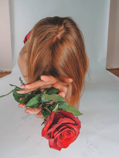 A young brown-haired woman with an attractive appearance in a red negligee with a red rose flower in her hands on a light wall background, close-up, the concept of the holidays of St. Valentine,March 8,signs of attention, romantic dates,female beauty - Φωτογραφία, εικόνα