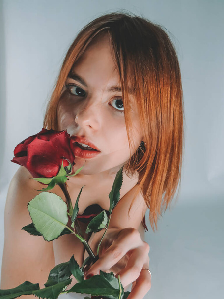 A young brown-haired woman with an attractive appearance in a red negligee with a red rose flower in her hands on a light wall background, close-up, the concept of the holidays of St. Valentine, March 8, romantic dates, female beauty - Zdjęcie, obraz