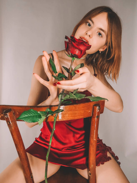 Attractive slender young red-haired woman on an old wooden chair in a red negligee enjoys the fragrance of a large red rose with water drops on the petals - Foto, Bild