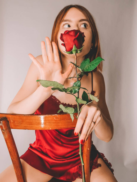 Atrakcyjna, szczupła, młoda rudowłosa kobieta na starym drewnianym krześle w czerwonym negliżu cieszy się zapachem dużej czerwonej róży z kroplami wody na płatkach. - Zdjęcie, obraz