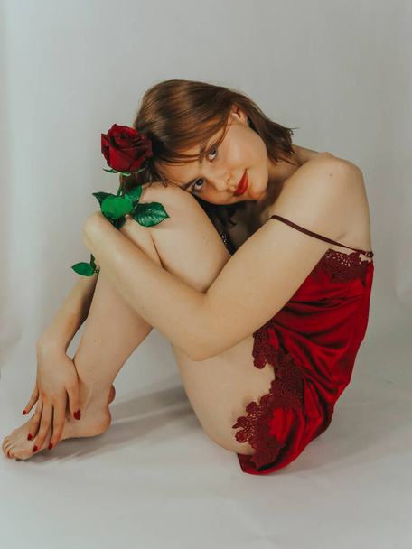 A young brown-haired woman with an attractive appearance in a red negligee with a red rose flower in her hands on a light wall background, close-up, the concept of the holidays of St. Valentine, March 8, congratulations and gifts to women - Foto, immagini