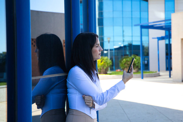 jonge, mooie, donkerharige vrouw raadpleegt haar mobiele telefoon bij de ingang van het gebouw waar ze als kantoormedewerker werkt. Begrip "werkende vrouw" en "zakenvrouw". - Foto, afbeelding