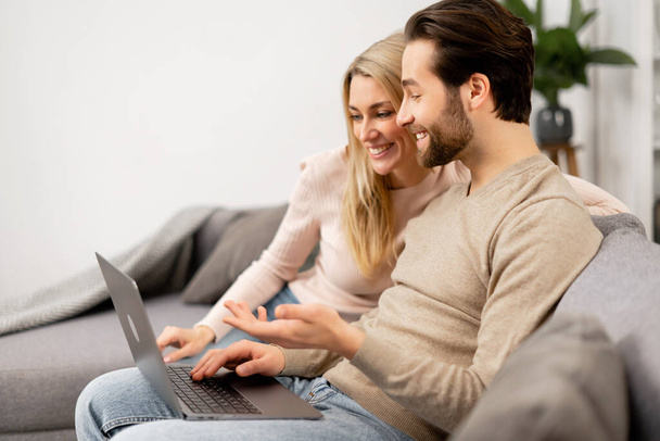 Happy girlfriend and her boyfriend looking at the laptop screen while sitting on the sofa, watching movies. Caucasian couple in love spending leisure time - Foto, immagini