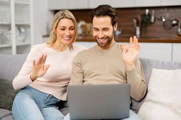 友達とのビデオ通話。ビデオ接続のためにラップトップを使用して陽気な夫婦。妻と夫は快適なソファに座っている間に笑顔でウェブカメラに向かって手を振っています - 写真・画像