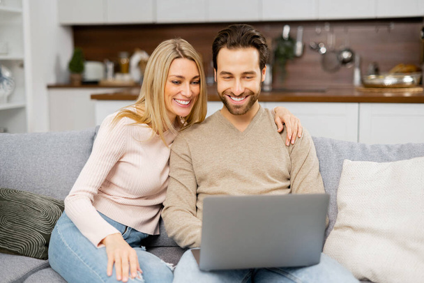 Blonde woman and man looking at the laptop screen while sitting on the sofa and watching comedy movies. Overjoyed couple in love spending leisure time online - Foto, Imagem