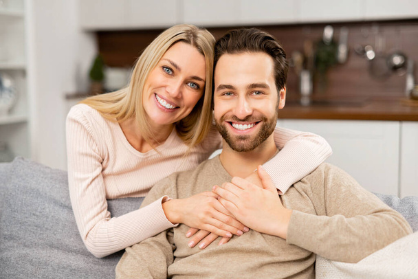 Portrait of lovely couple at home. Handsome man sitting on the comfortable couch, beautiful blonde woman hugging his from the back - Foto, afbeelding