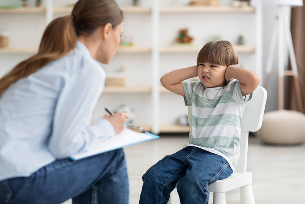 Ammattimainen psykoterapeutti yrittää puhua vaikea lapsi, äreä pieni poika peittää korvat ja rypistynyt nenä - Valokuva, kuva
