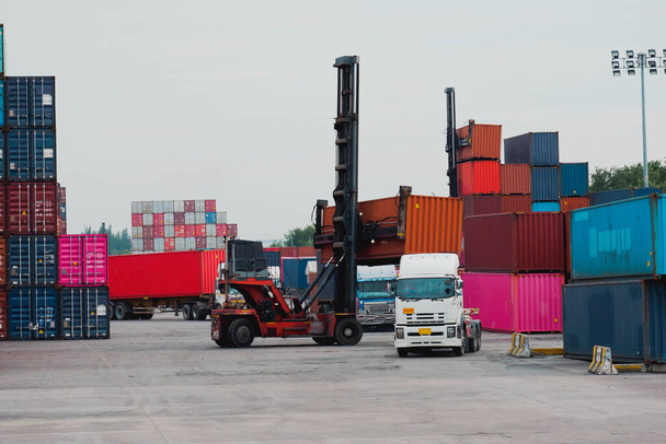 Вилочный погрузчик поднимает грузовой контейнер на судоходной верфи или в доках на фоне восходящего неба со штабелем грузовых контейнеров на заднем плане для транспортировки импорта, экспорта и логистической промышленной концепции
 - Фото, изображение