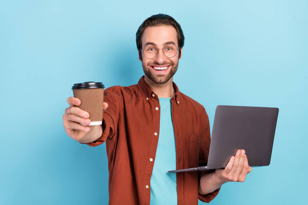 Photo of cute brunet young guy hold laptop give coffee wear spectacles bown shirt isolated on blue color background - Photo, Image