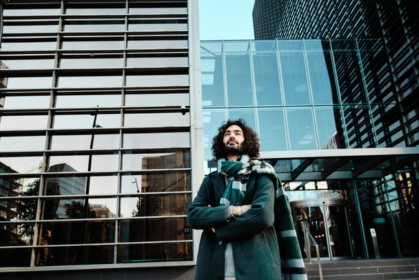 Een man kijkt weg terwijl hij poseert met zijn armen gekruist tegen een kantoorgebouw in het financiële district.. - Foto, afbeelding
