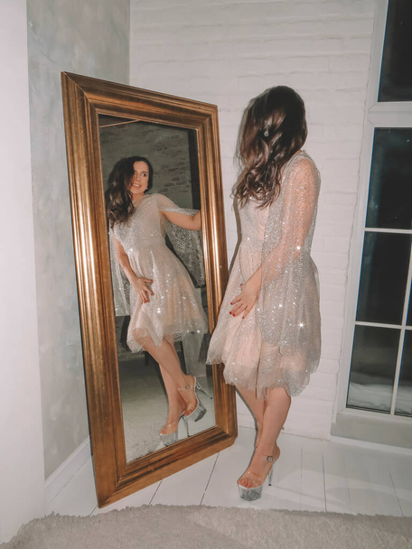 A young black-haired bride tries on a white silk dress in front of a mirror, the concept of a holiday, a wedding, emotions of love and happiness - Фото, изображение
