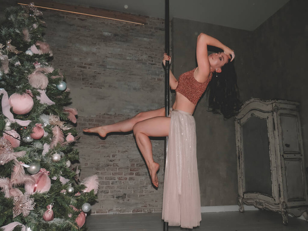 A young attractive black-haired stripper woman performs exercises on a pole near a Christmas tree, the concept of sports and fitness, acrobatics on a pole, striptease dancing, beauty and sexuality of the female body, New Year's holidays - Foto, immagini