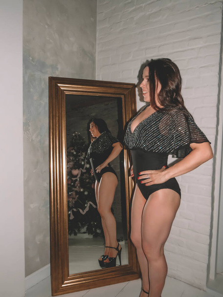 Attractive black-haired young female gymnast in a black bodysuit with a slim athletic body in front of a mirror, the concept of sports and fitness, beauty, flexibility, plasticity of the body, a healthy active lifestyle - 写真・画像