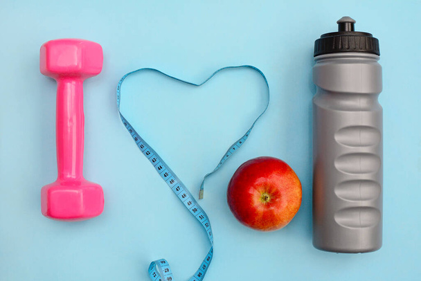 The concept of fitness and healthy lifestyle. Dumbbells, bottle, apple and ribbon on blue background, top view - Zdjęcie, obraz