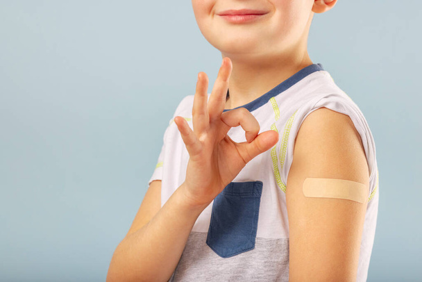 子供の予防接種。ワクチン注射後、幸せなワクチン接種された子供の男の子は元気にジェスチャーし、接着剤包帯で腕を見せています。子供とcovid-19予防、抗ウイルス予防接種. - 写真・画像