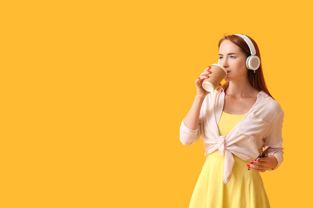 Young redhead woman in headphones with mobile phone drinking coffee on yellow background - Photo, image