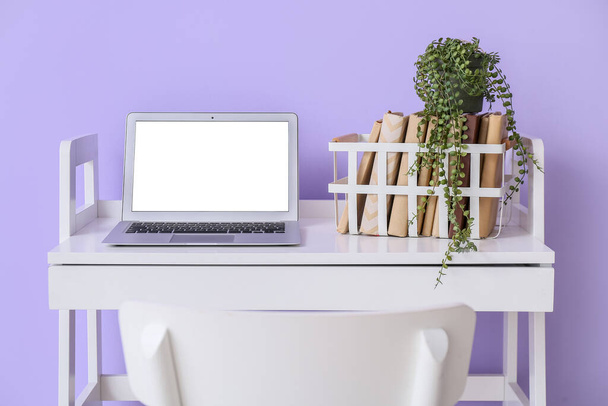 Moderne werkplaats met laptop, boeken en kamerplant nabij violette muur - Foto, afbeelding