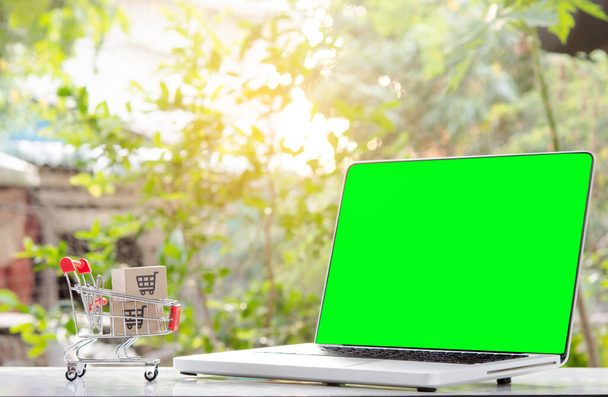 Ostoksia verkossa. pahvilaatikko, jossa on ostoskorin logo vaunussa ja kannettava vihreä näyttö. Ostospalvelu verkossa. tarjoaa kotiinkuljetus - Valokuva, kuva