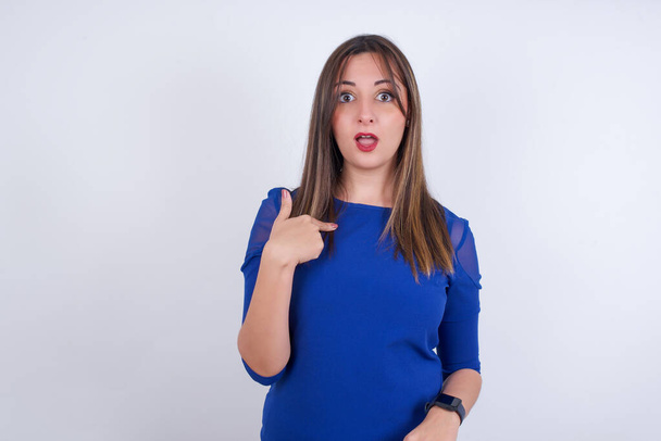 Mujer árabe joven vistiendo vestido azul sobre blanco backgtound estar en estupor sorprendido, tiene expresión asombrada señalándose con el dedo diciendo: ¿Quién me? - Foto, imagen