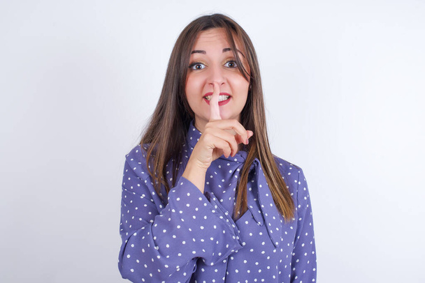 Glimlachende jonge Arabische vrouw draagt polka-dot kleding over witte rug maakt een stil gebaar, houdt de wijsvinger boven de lippen verborgen. Wees stil, alsjeblieft.. - Foto, afbeelding