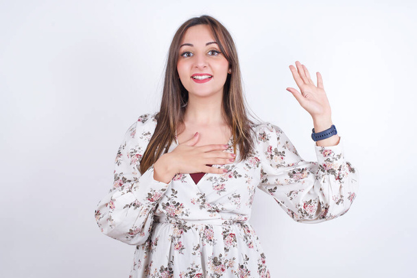 Promets-moi de ne pas regretter. Portrait de sincère jeune femme arabe portant une chemise florale sur fond blanc levant un bras et tenant la main sur le cœur comme donner serment, dire la vérité, vous voulez croire. - Photo, image