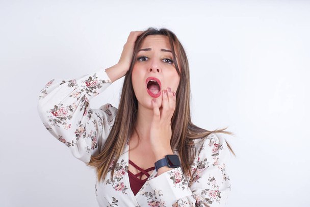 Молодая мрачная арабская женщина в цветочной рубашке на белом фоне, прячущая лицо с опухшими руками и плачущими, стоя расстроенная и подавленная жалобами на проблемы с работой. - Фото, изображение