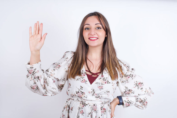 Jeune femme arabe portant une chemise florale sur fond blanc renonçant à dire bonjour ou au revoir heureux et souriant, geste de bienvenue amical. - Photo, image