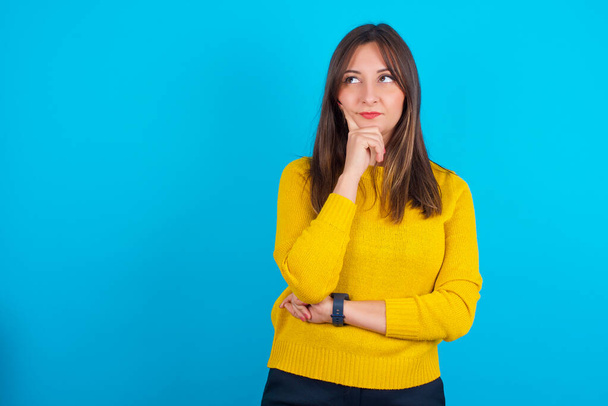 Thoughtful Young arab woman wearing yellow knitted sweater over blue backgtound holds chin and looks away pensively makes up great plan - Фото, изображение