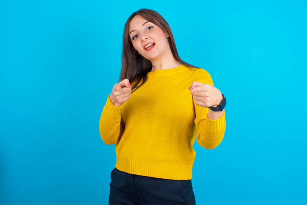 Mujer árabe joven con suéter de punto amarillo sobre backgtound azul dirige los dedos a la cámara selecciona a alguien. Te recomiendo. La mejor opción - Foto, imagen