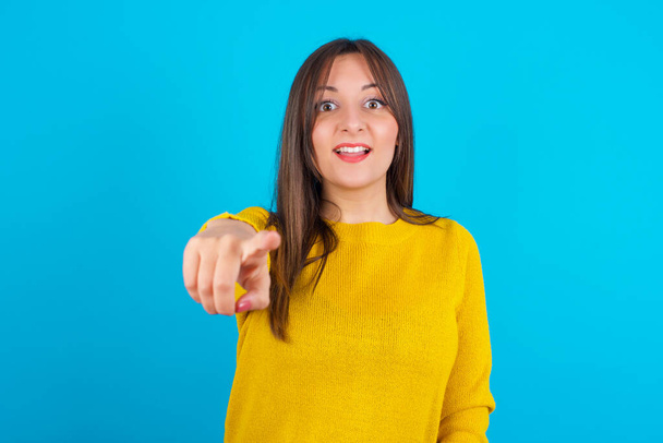 Opgewonden positief Jonge arabische vrouw met een geel gebreide trui over een blauwe ruggengraat wijst naar je wijsvinger, ziet iets heel grappigs. Wauw, geweldig. - Foto, afbeelding