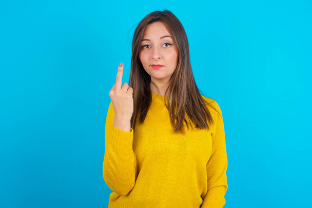 Young arab woman wearing yellow knitted sweater over blue backgtound shows middle finger bad sign asks not to bother. Provocation and rude attitude. - Фото, зображення