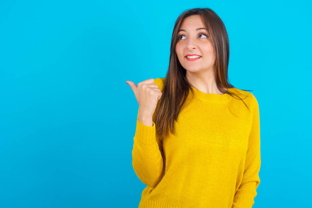 Νεαρή αραβίδα γυναίκα φορώντας κίτρινο πλεκτό πουλόβερ πάνω από το μπλε backgtound σημεία μακριά και δίνει συμβουλές αποδεικνύει διαφήμιση - Φωτογραφία, εικόνα