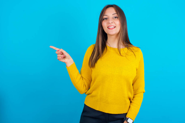 Молодая арабская женщина в желтом трикотажном свитере над синим задним числом смеется счастливо указывает прочь на пустое место демонстрирует торговый скидки предложение, взволнованный хорошей новостью или неожиданной продажи. - Фото, изображение