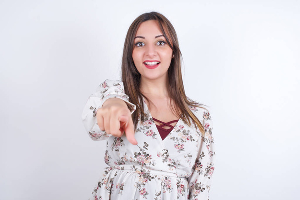 Возбужденная положительная женщина в цветочном платье на белом фоне указывает указательным пальцем прямо на вас, видит что-то очень смешное. Wow, amazing - Фото, изображение