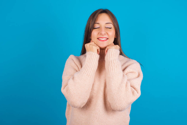 Young arab woman wearing knitted sweater over blue backgtound grins joyfully, imagines something pleasant, copy space. Pleasant emotions concept. - Photo, Image