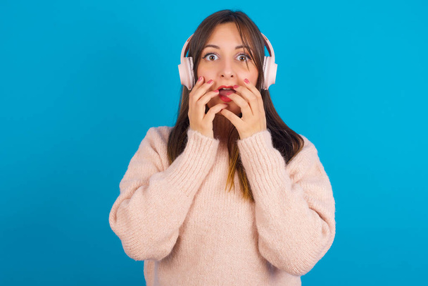 Šokovaný mladý arab žena na sobě pletený svetr přes modrý backgtound zírá ustrašený na kameru udržuje ústa široce otevřené nosí bezdrátové stereo sluchátka na uších - Fotografie, Obrázek