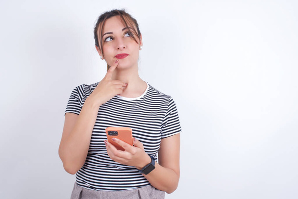 молодая красивая белая женщина в футболке о чем-то глубоко думает, пользуется современным мобильным телефоном, пытается придумать хорошее сообщение, держит указательный палец у губ. - Фото, изображение