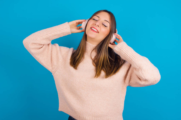 Jonge arabische vrouw draagt gebreide trui over blauwe backgtound glimlacht over het algemeen voelt erg blij luistert favoriete muziek track via draadloze hoofdtelefoon sluit ogen. - Foto, afbeelding