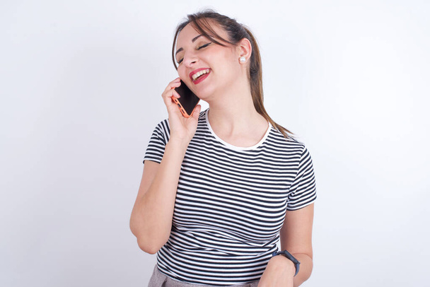 Divertido joven hermosa mujer caucásica vistiendo camiseta ríe felizmente, tiene conversación telefónica, siendo divertido por amigo, cierra los ojos. - Foto, imagen
