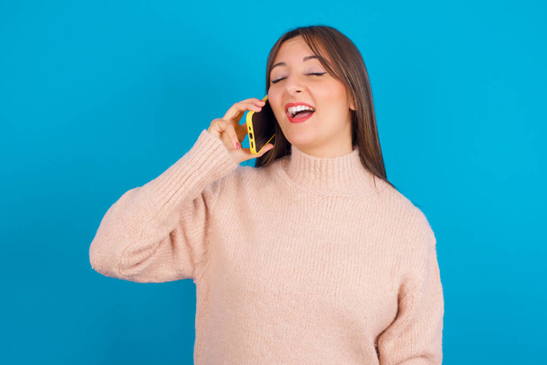 Overemotive szczęśliwy młody arabski kobieta w dzianiny sweter na niebieski backgtound śmieje się pozytywnie słyszy zabawną historię od przyjaciela podczas rozmowy telefonicznej - Zdjęcie, obraz