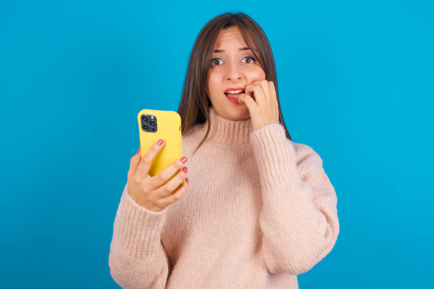 Vrees grappig Jonge arabische vrouw draagt gebreide trui over blauwe achterzijde telefoon en bijtende nagels - Foto, afbeelding