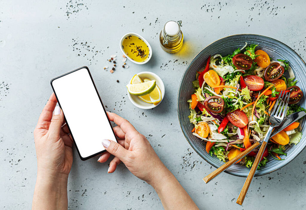 Smartfon w rękach kobiety obok świeżo ubarwionej sałatki warzywnej. Używanie telefonu podczas lunchu. Restauracja żywności - menu online lub koncepcji przeglądu. Wolna przestrzeń do kopiowania. - Zdjęcie, obraz