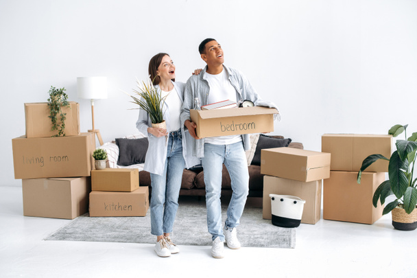 Moving to a new home. Excited couple in love, husband and wife of different nationalities, moved to their new home, guy holds a box with things, girl a flowerpot, enthusiastically inspecting new house - Photo, Image