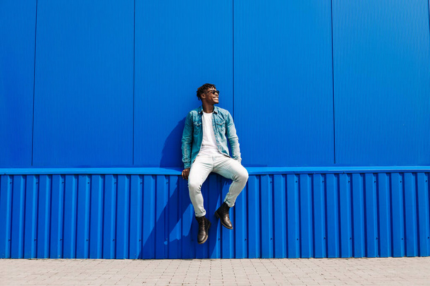 Handsome african male fashion model in sunglasses smiling and posing while standing near blue wall outdoors. Stylish curly african man smiling over isolated blue background - Фото, зображення