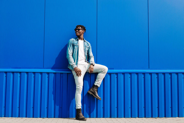 Handsome african male fashion model in sunglasses smiling and posing while standing near blue wall outdoors. Stylish curly african man smiling over isolated blue background - Fotoğraf, Görsel