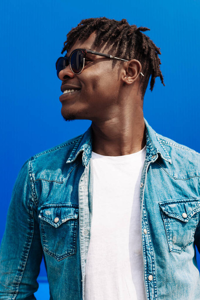 Jóképű, divatos afro-amerikai hipszter férfi stílusos napszemüvegben, kék fal mellett pózol. Jóképű fiatal fekete férfi pózol a stúdióban - Fotó, kép