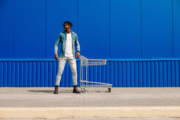 Handsome African American man shopping in push cart supermarket. Side view of a cheerful young man, with a shopping trolley, over a blue wall with copy space. The concept of consumerism, shopping - Foto, afbeelding