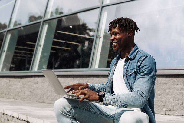 Close-up of an African American student using his laptop outdoors while working in a modern business center, modern buildings in the background - Photo, Image