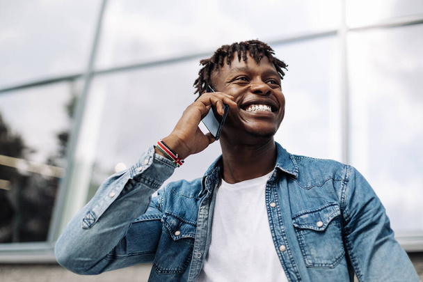Image of a handsome smiling African man talking on his cell phone while walking on a city street. Dark-skinned man with a cell phone having a conversation on a mobile - Foto, imagen